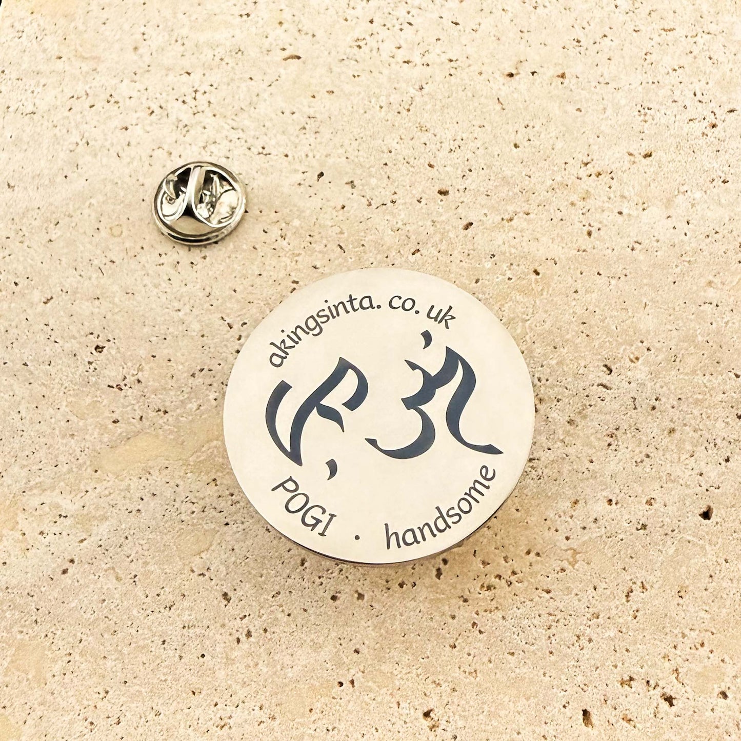 An Aking Sinta product photograph showcasing a circular silver lapel pin with its butterfly clutch back on a textured light stone surface. The polished pin is custom-engraved with the Aking Sinta brand website akingsinta.co.uk along the top edge, traditional Baybayin script characters in the center, and the Tagalog word "POGI" with its English translation "handsome" at the bottom