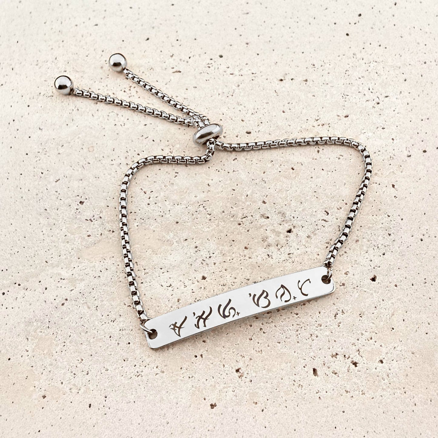 A silver Aking Sinta box chain bracelet featuring a bespoke engraving. The flat silver bar is etched with ancient Filipino Baybayin script. The jewelry includes an adjustable bolo-style slider clasp with spherical end beads, displayed on a light natural stone surface