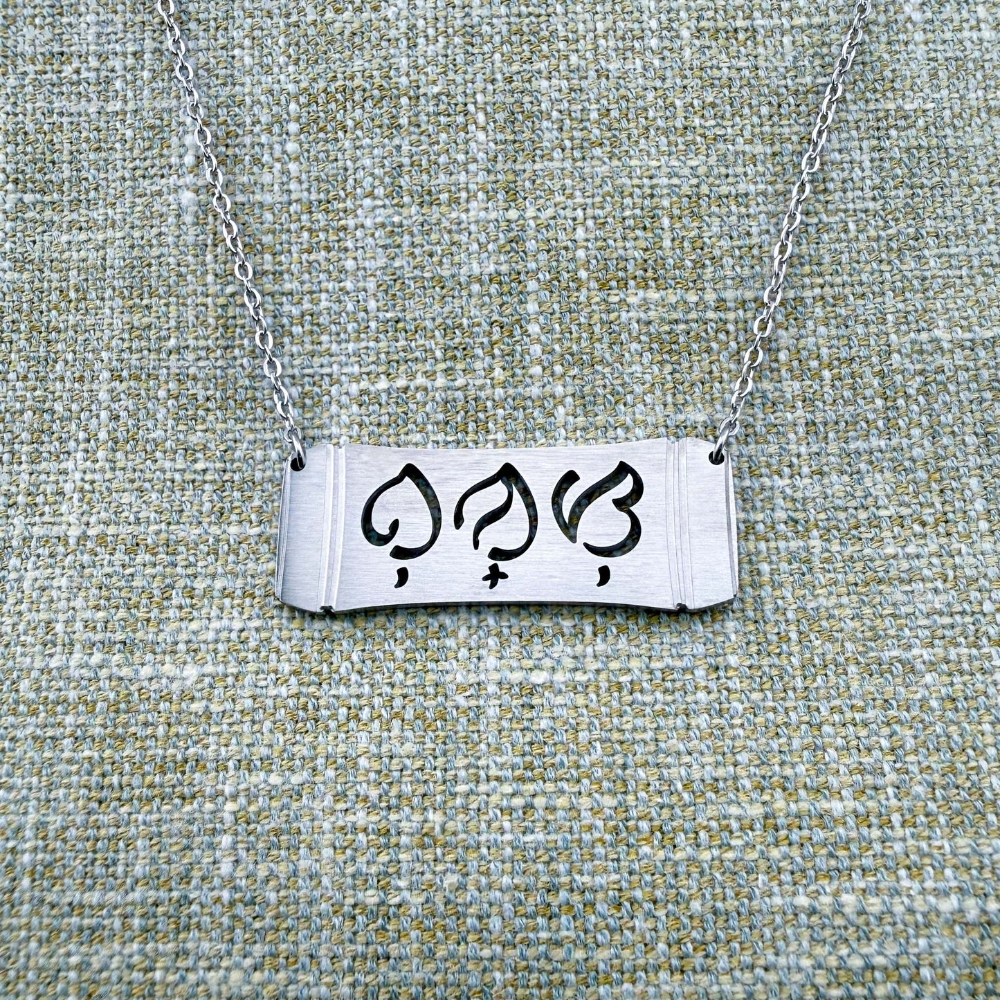 An Aking Sinta silver horizontal bar necklace displayed on a textured green-gray linen background. The matte silver pendant features a curved bamboo frame silhouette with the word "Bunso" (Youngest Child) precisely cut out in script-like traditional Baybayin characters, connected to a fine silver link chain.