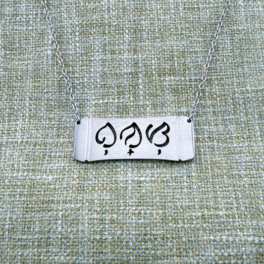 An Aking Sinta silver horizontal bar necklace displayed on a textured green-gray linen background. The matte silver pendant features a curved bamboo frame silhouette with the word "Bunso" (Youngest Child) precisely cut out in script-like traditional Baybayin characters, connected to a fine silver link chain.