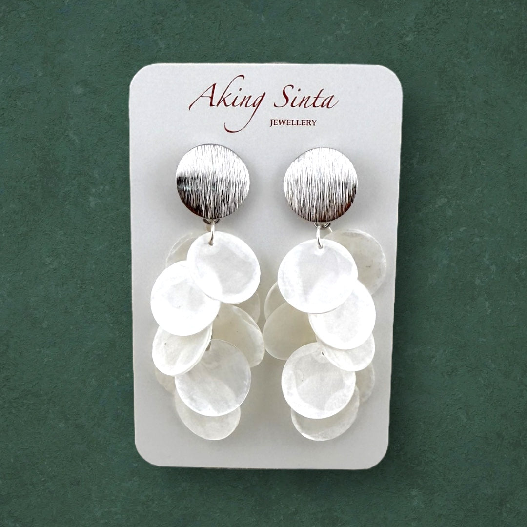 A pair of Aking Sinta silver Cascading Capiz Shell Earrings with brushed finish studs; the earrings are mounted on a white cardboard display holder with "Aking Sinta" branding at the top, set against a dark green background.