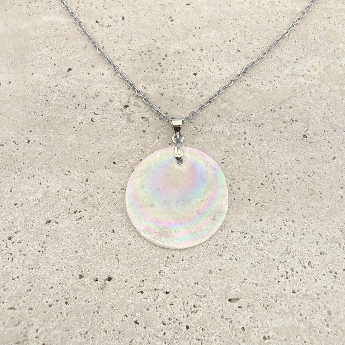 A silver Aking Sinta necklace featuring a large, circular, iridescent Capiz shell pendant. The natural shell disc showcases beautiful shimmering, rainbow-like hues, suspended from a polished silver bail and a delicate silver cable chain on a light, textured natural stone surface