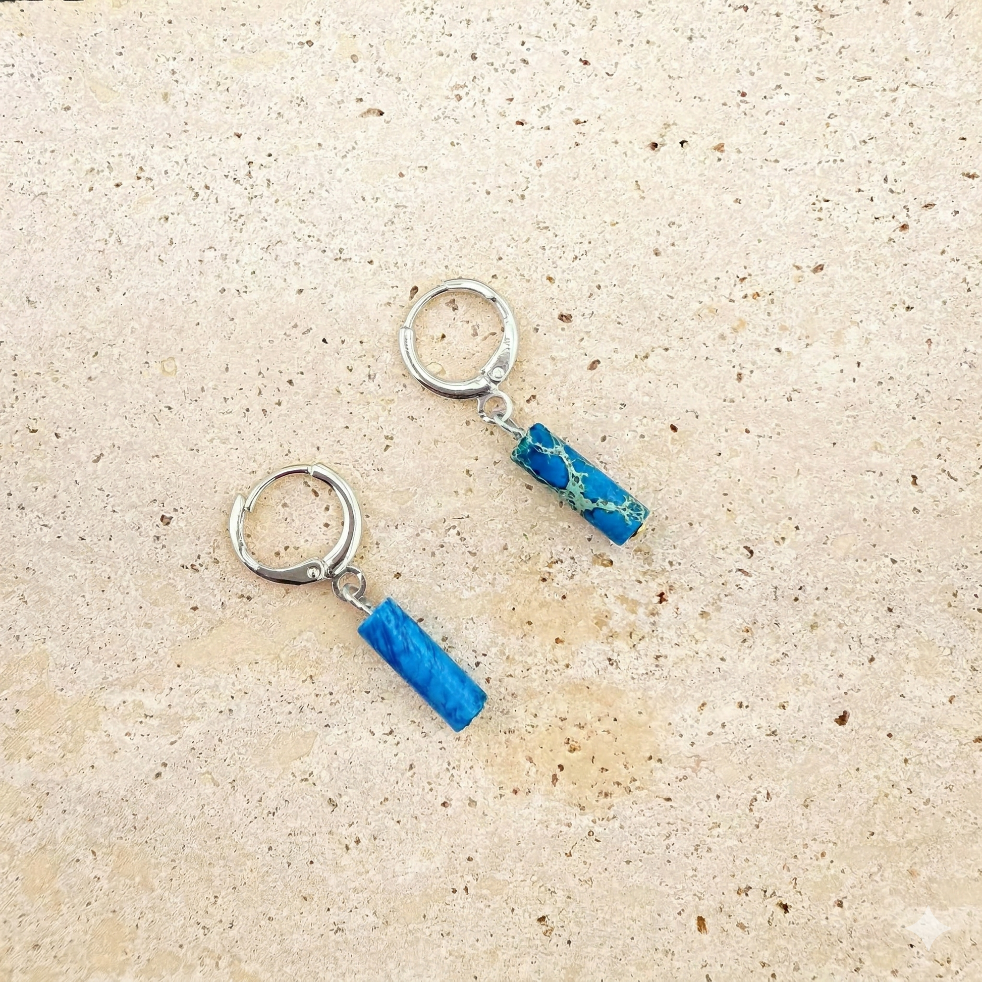 Silver-toned stainless steel huggie earrings with circular ocean blue Imperial Jasper charms on a white marble background.