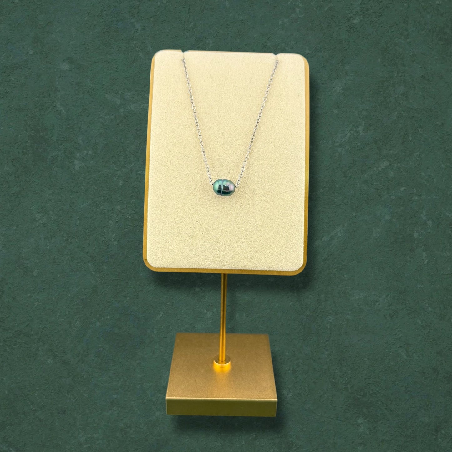 A minimalist Aking Sinta dainty silver chain necklace featuring a single, iridescent dark pearl pendant. The necklace is elegantly showcased on a cream-colored velvet stand with a gold base, set against a rich, dark green textured background, highlighting its sophisticated and refined design