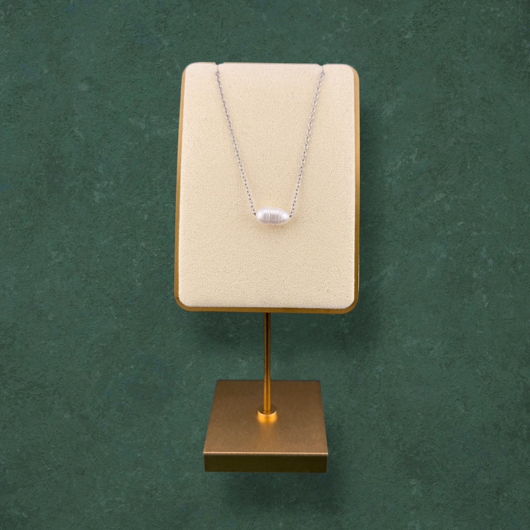 A minimalist Aking Sinta dainty silver chain necklace featuring a single, lustrous white pearl pendant. The necklace is elegantly showcased on a cream-colored velvet stand with a gold base, set against a rich, dark green textured background, highlighting its sophisticated and refined design