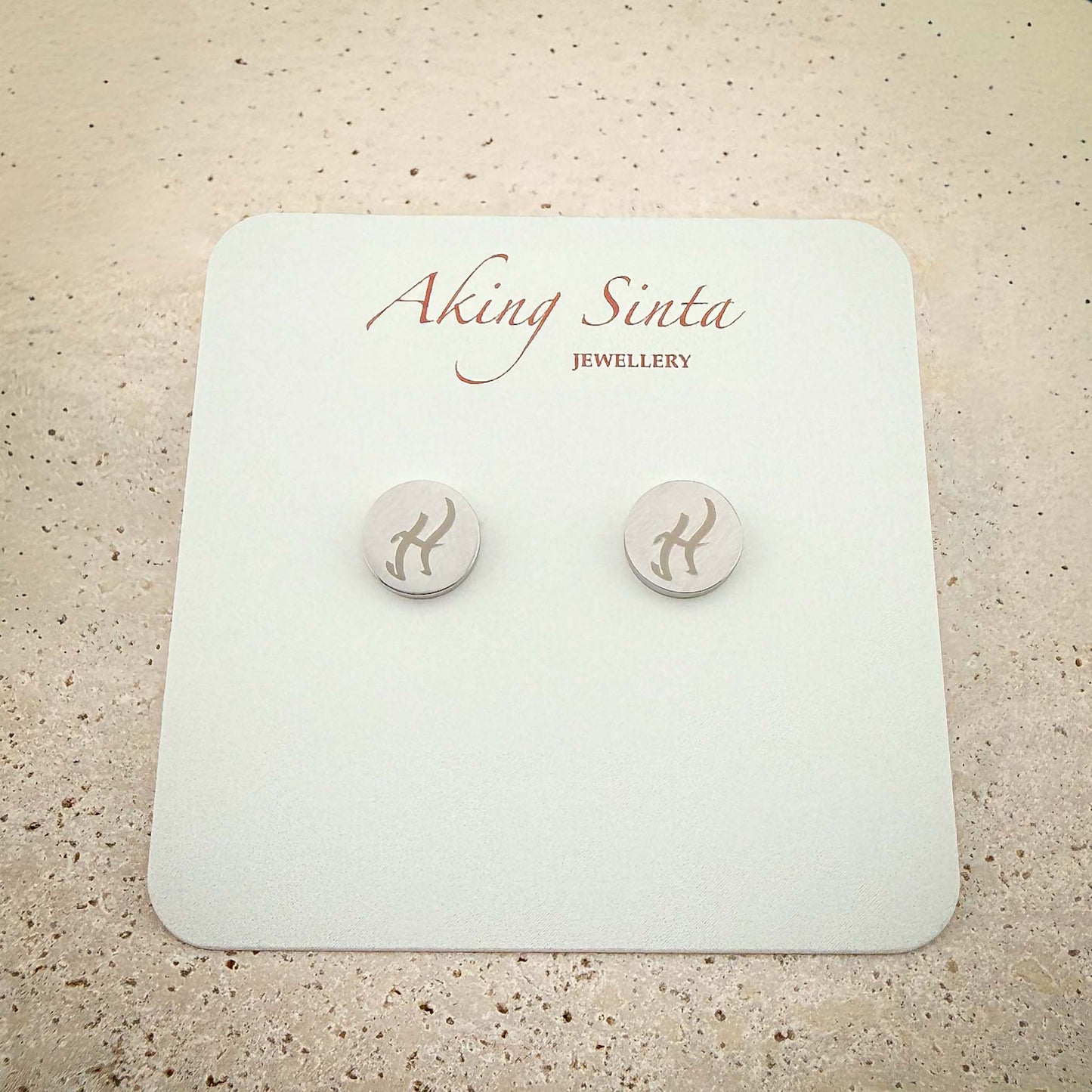 A pair of silver dainty stud earrings from Aking Sinta Jewellery featuring bespoke engravings. The small circular silver studs are etched with ancient Filipino Baybayin script and presented on a white branded display card, set against a light-colored natural stone background