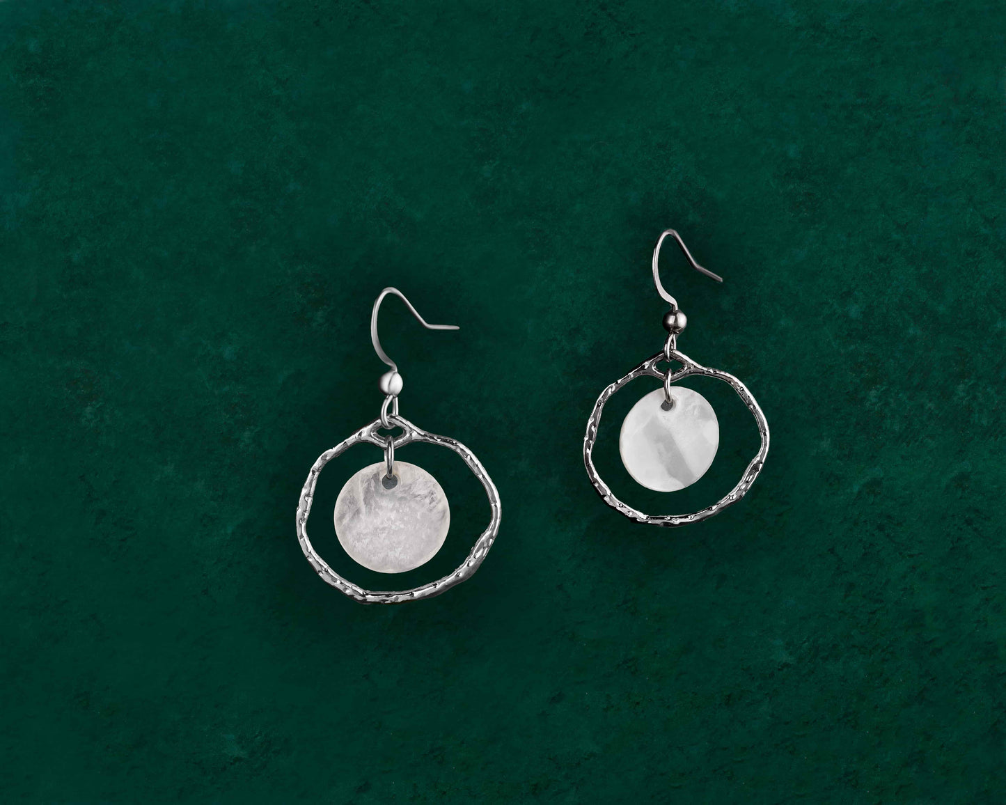 A pair of Aking Sinta silver Floating Capiz Shell Hook Earrings displayed on a dark green background; featuring minimalist polished silver ear hooks with a single, round, translucent white Capiz shell disk hanging freely.