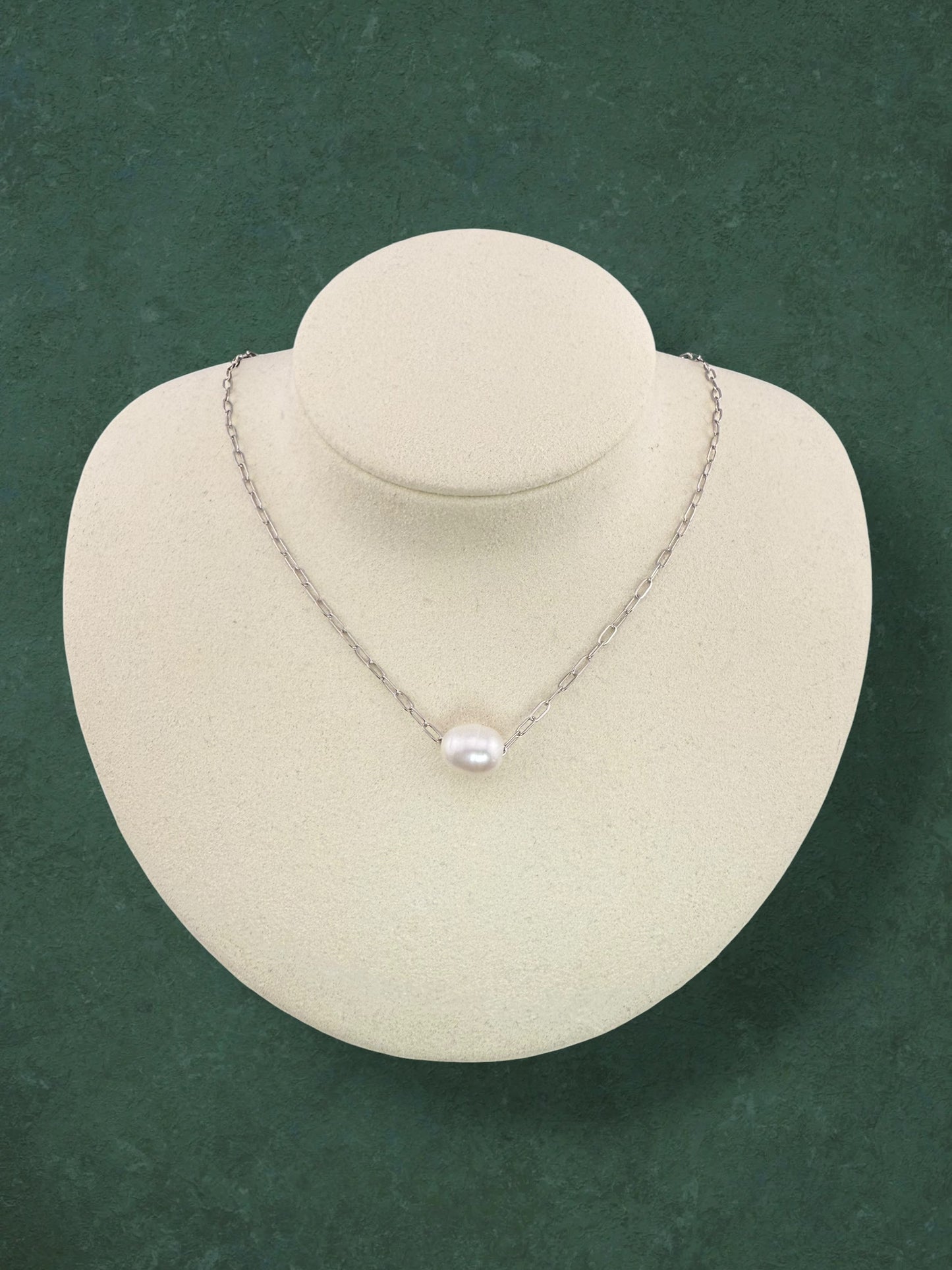 An Aking Sinta silver paperclip chain necklace with a single floating white baroque pearl. This minimalist jewelry piece is showcased on a white velvet display bust against a textured dark green background.