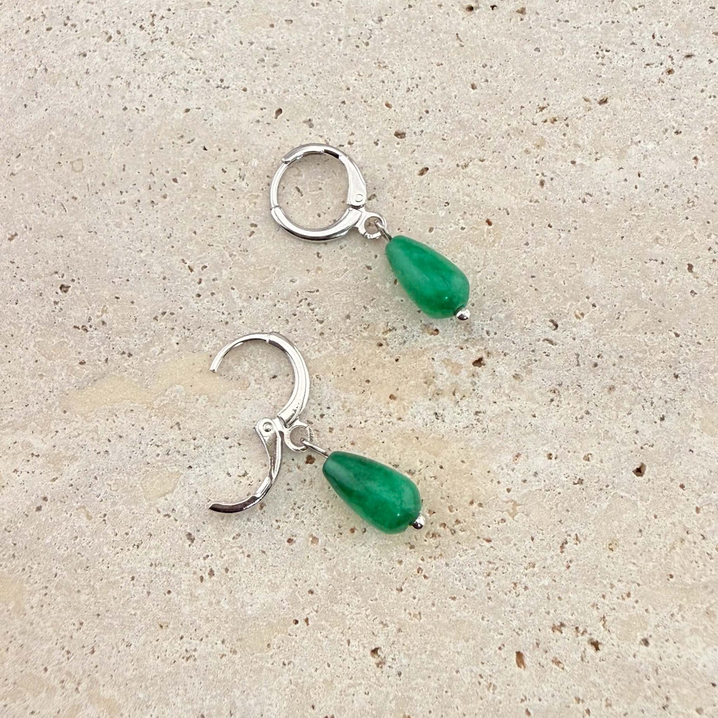A pair of Aking Sinta silver huggie earrings with polished green jade teardrop pendants. One earring is unlatched to demonstrate the hinge closure, shown on a light-colored natural stone background.
