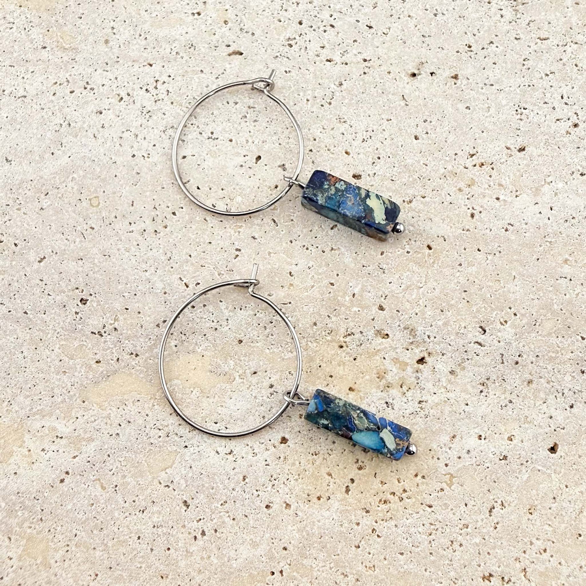 A pair of Aking Sinta silver wire hoop earrings featuring blue imperial jasper cuboid gemstone pendants. The unique, marbled blue and earth-toned stone drops are displayed on a light sand-colored stone surface