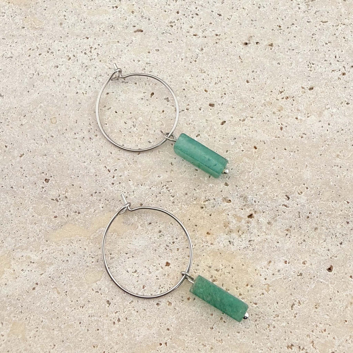 A pair of Aking Sinta silver wire hoop earrings featuring green aventurine cuboid gemstone pendants. The minimalist silver hoops and natural green stone drops are displayed on a light sand-colored stone surface.