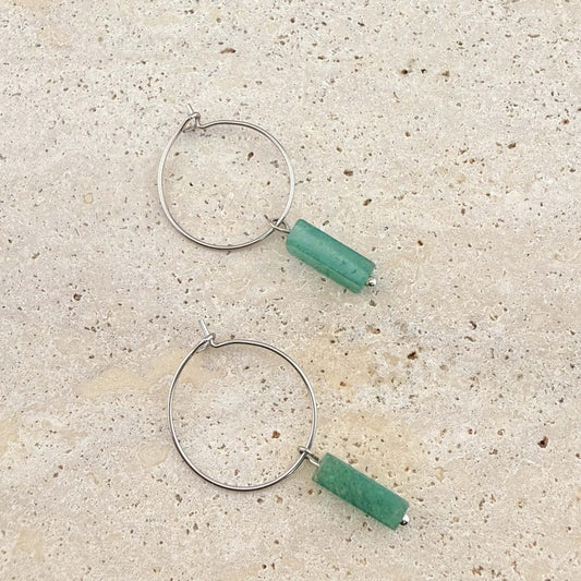 A pair of Aking Sinta silver wire hoop earrings featuring green aventurine cuboid gemstone pendants. The minimalist silver hoops and natural green stone drops are displayed on a light sand-colored stone surface.