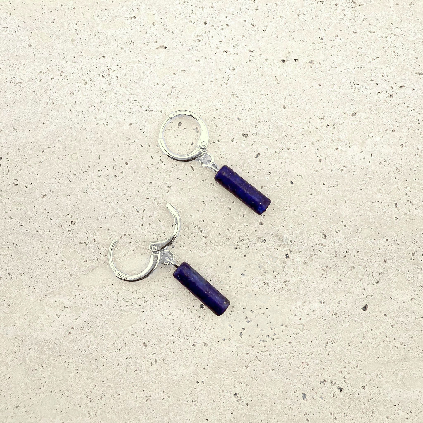 A pair of Aking Sinta silver huggie hoop earrings featuring polished deep blue Lapis Lazuli column drops. One hoop is shown closed and the other open to reveal the secure hinge clasp mechanism. The earrings are laid out on a light, textured natural stone surface, highlighting the rich cobalt blue of the gemstones against the bright silver