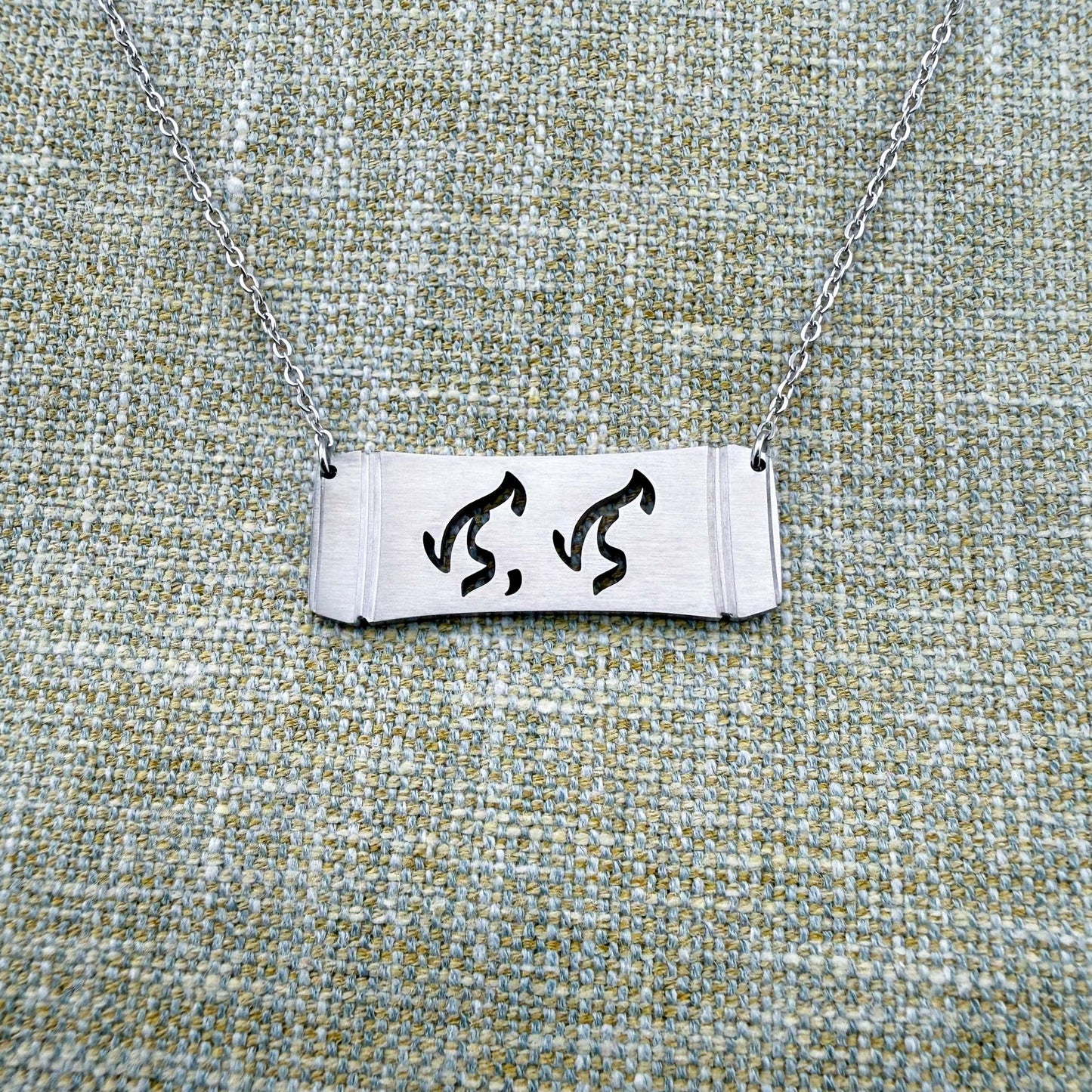 An Aking Sinta silver horizontal bar necklace centered on a textured green-gray linen background. The matte silver pendant features a curved bamboo frame silhouette with the word "Lola" (Grandmother) elegantly cut out in traditional script-like Baybayin characters, suspended by a fine silver link chain.