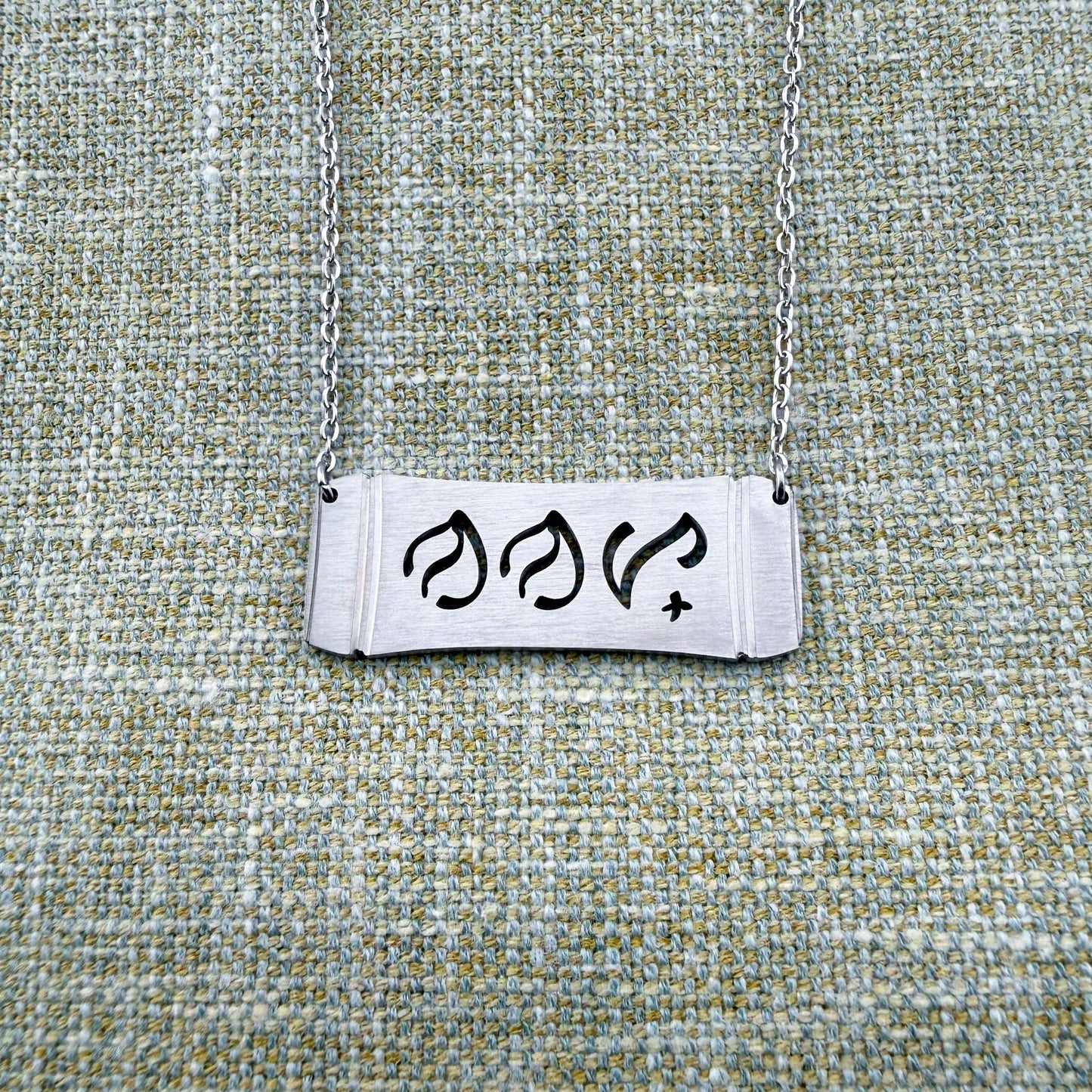 An Aking Sinta silver horizontal bar necklace displayed on a textured green-gray linen fabric background. The matte silver pendant features a stylized bamboo frame silhouette with the word "Nanay" (Mother) cleanly cut out in traditional Baybayin script characters, suspended from a fine silver link chain.