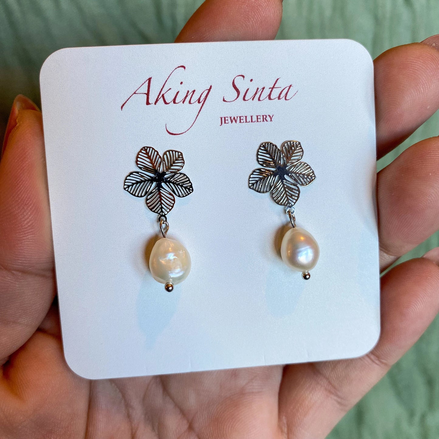 A pair of silver-tone Aking Sinta statement earrings presented on a branded white display card. Each earring features an intricate filigree Sampaguita flower stud with a dangling, organic white baroque pearl drop, held in a hand against a soft green background