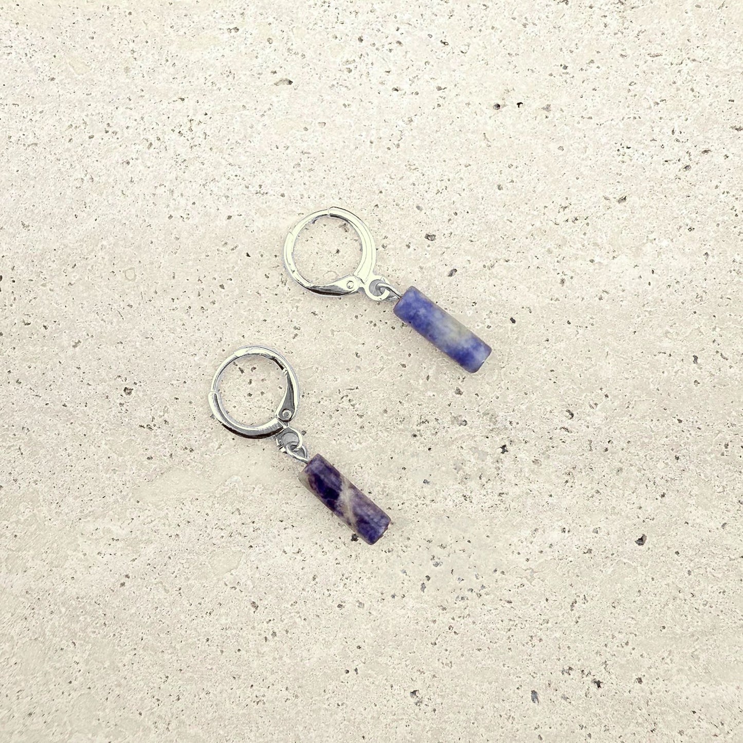 A pair of Aking Sinta silver huggie hoop earrings featuring blue sodalite column charms. The natural gemstone cylinders display unique marbled patterns of indigo and white, suspended from polished silver-toned hoops on a light natural stone surface.