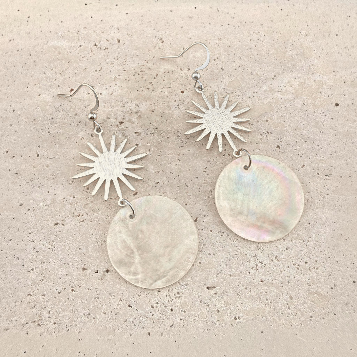 A pair of Aking Sinta silver drop earrings featuring a radiant, brushed silver sun motif attached to circular, iridescent white Capiz shell pendants. The shimmering, natural shell discs are suspended from the suns and displayed elegantly on a light, textured stone surface