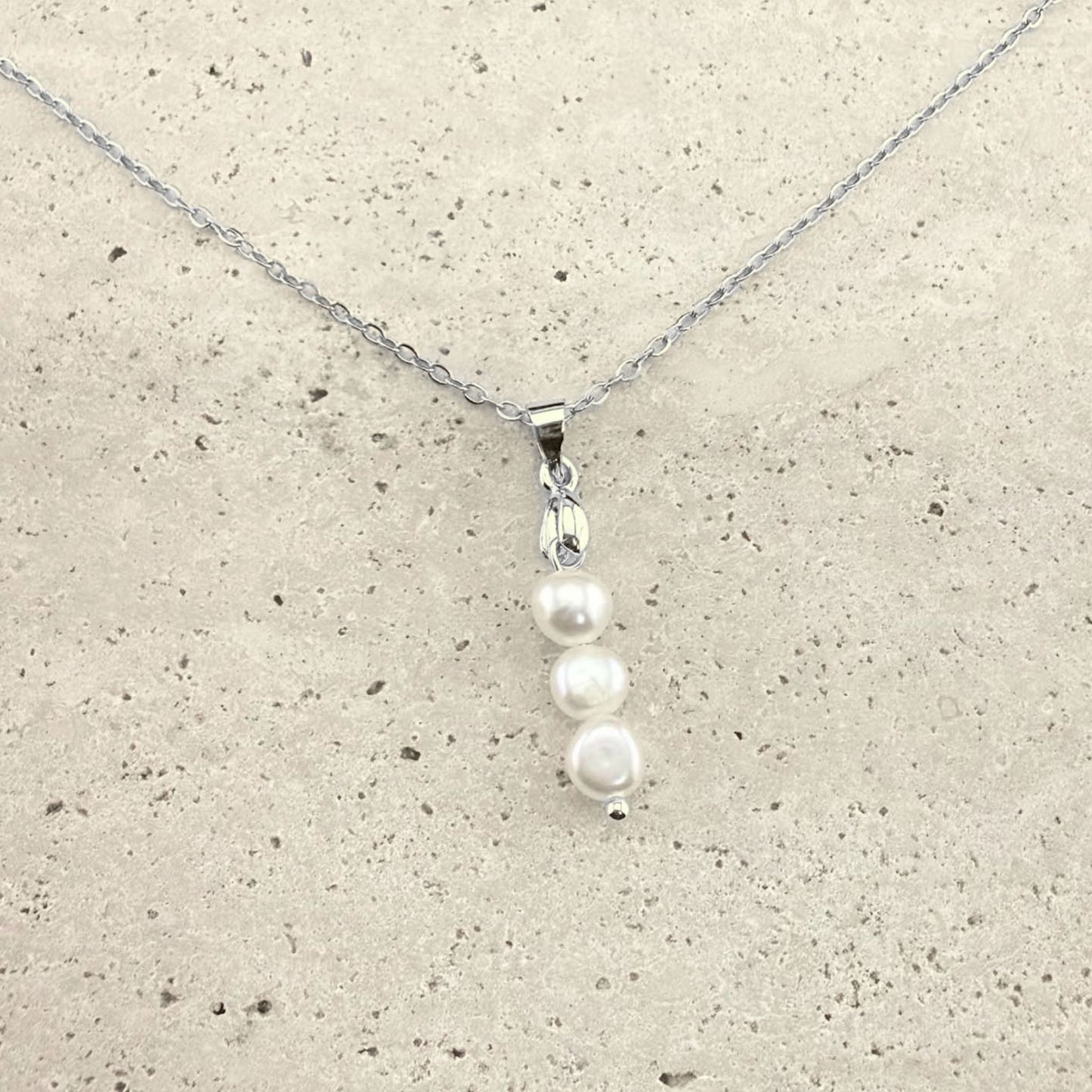 A silver Aking Sinta necklace featuring a delicate pendant composed of three white, organic-shaped baroque pearls stacked vertically. The pearls are suspended from a polished silver bail and a fine silver cable chain, presented on a light, textured natural stone surface