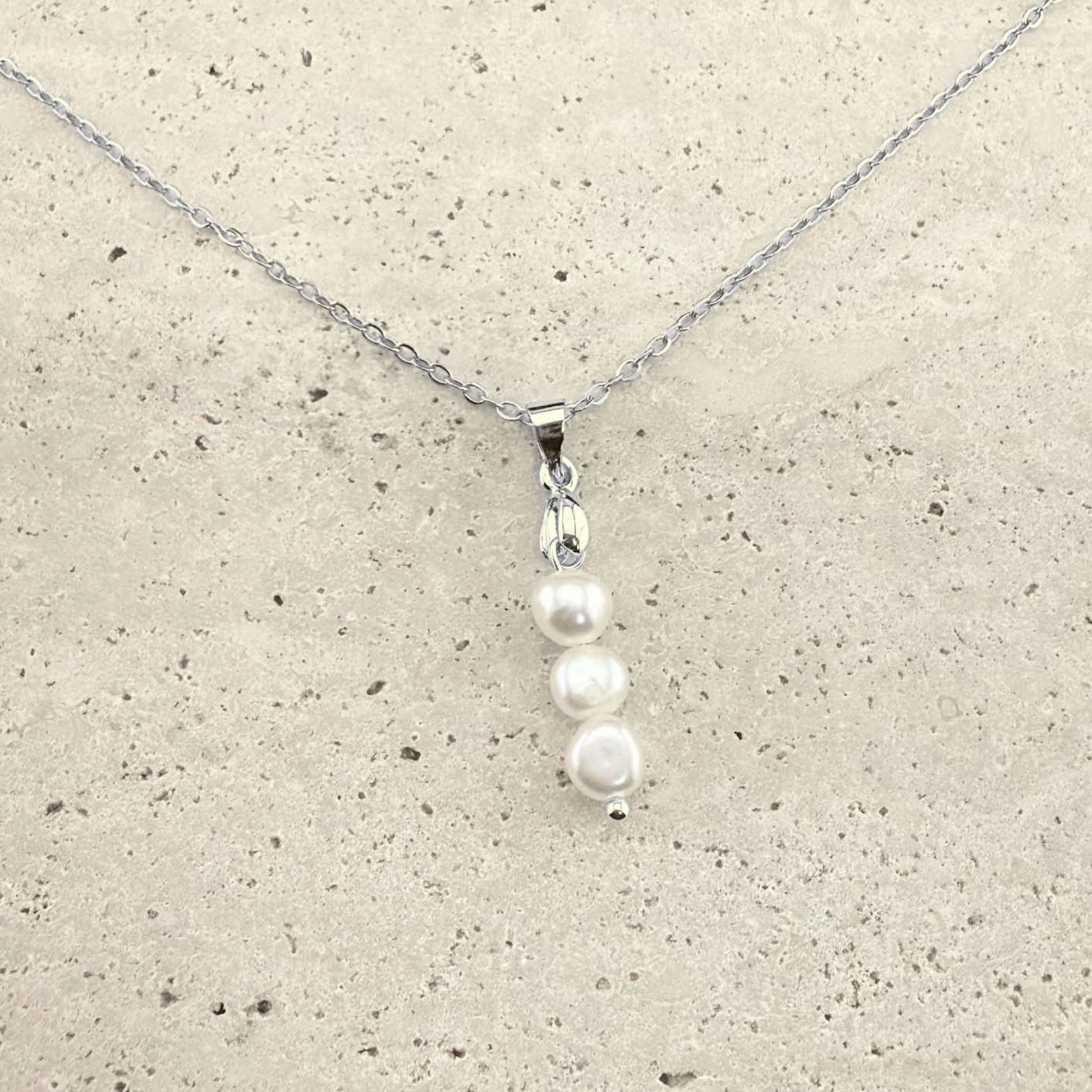 A silver Aking Sinta necklace featuring a delicate pendant composed of three white, organic-shaped baroque pearls stacked vertically. The pearls are suspended from a polished silver bail and a fine silver cable chain, presented on a light, textured natural stone surface
