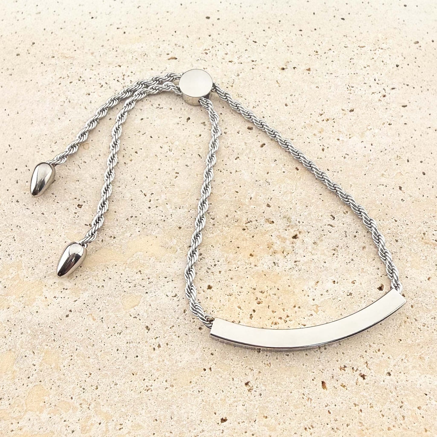 A silver Aking Sinta twisted rope chain bracelet with a polished, curved bar ready for bespoke engraving. The adjustable bolo-style bracelet features a circular slider clasp and elegant teardrop end charms, displayed on a light-colored natural stone surface.