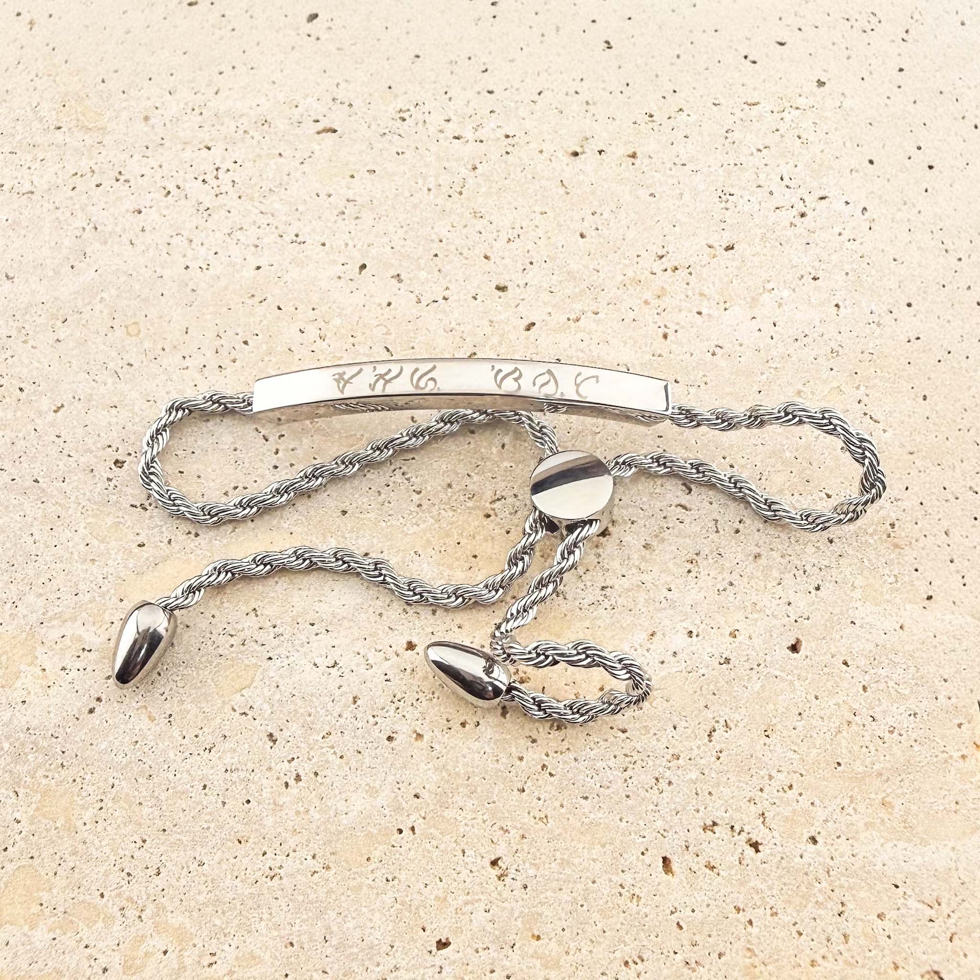 A silver Aking Sinta twisted rope chain bracelet with a bespoke engraving. The curved silver bar is etched with ancient Filipino Baybayin script and features an adjustable bolo-style slider clasp with teardrop-shaped end charms, displayed on a light natural stone surface