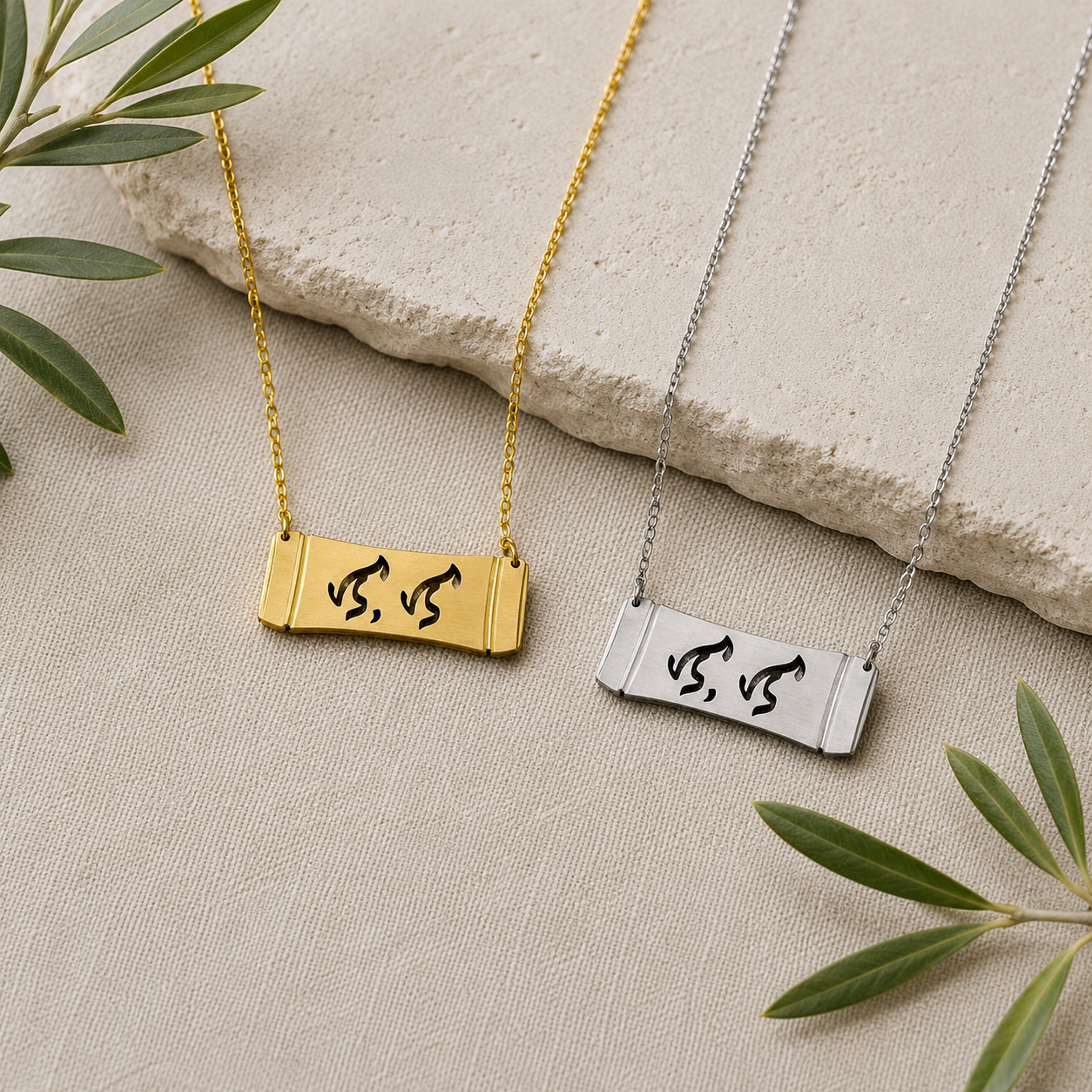A flat lay product photograph displaying two Aking Sinta horizontal bamboo bar necklaces in matte gold and silver finishes side by side. The parallel curved bar pendants feature the word "Lola" (Grandmother) elegantly cut out in traditional script-like Baybayin characters, styled on a neutral woven textile next to a textured stone slab and fresh olive branches