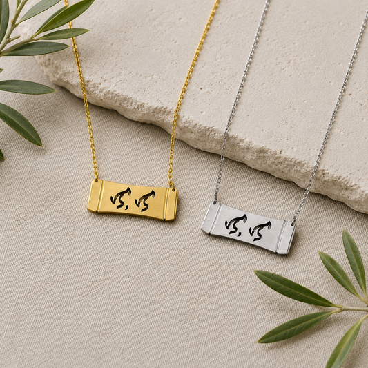 A flat lay product photograph displaying two Aking Sinta horizontal bamboo bar necklaces in matte gold and silver finishes side by side. The parallel curved bar pendants feature the word "Lola" (Grandmother) elegantly cut out in traditional script-like Baybayin characters, styled on a neutral woven textile next to a textured stone slab and fresh olive branches