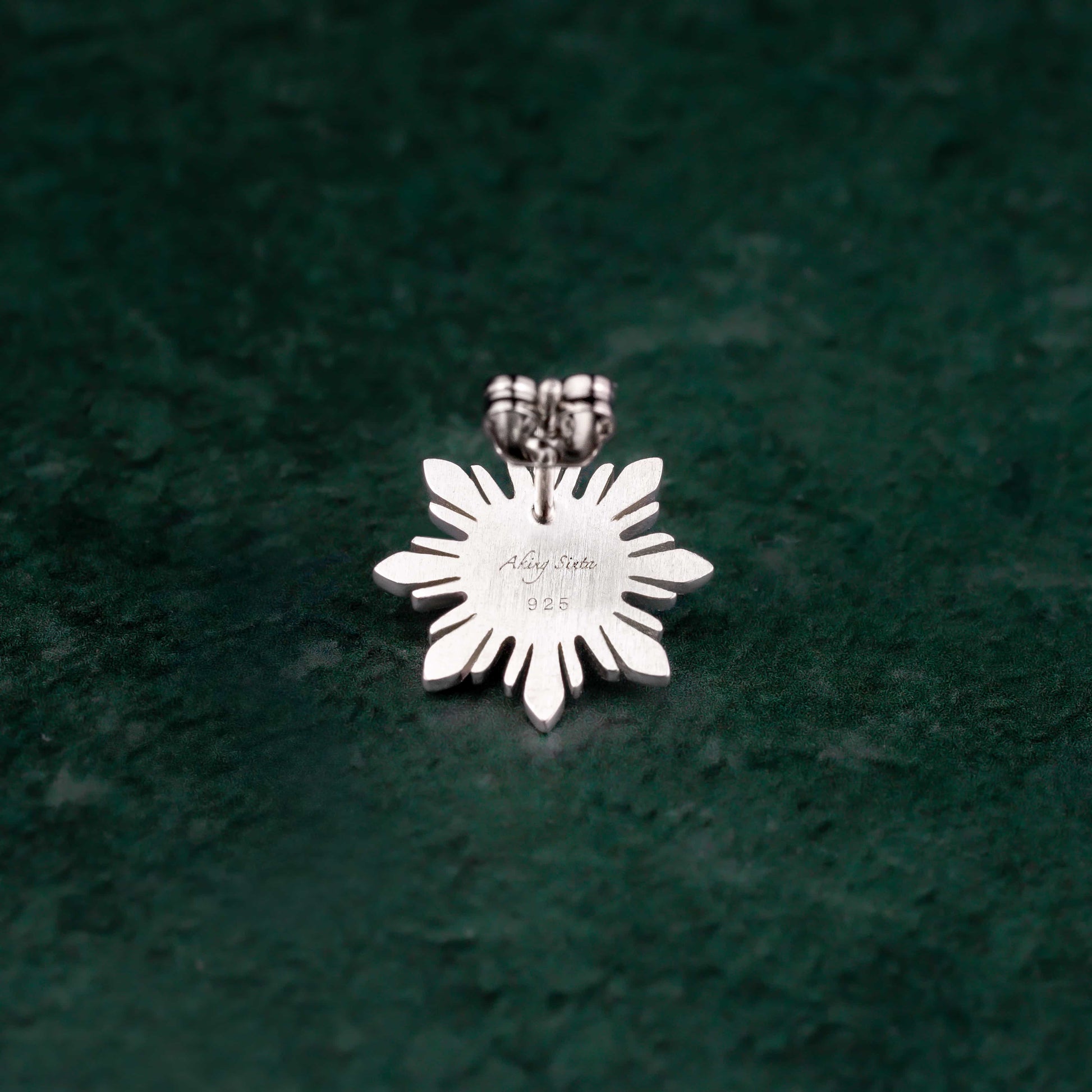 A single Aking Sinta Araw sterling silver stud earring featuring the detailed 8-ray sunburst design, positioned on a vibrant green background to showcase its fine polished finish