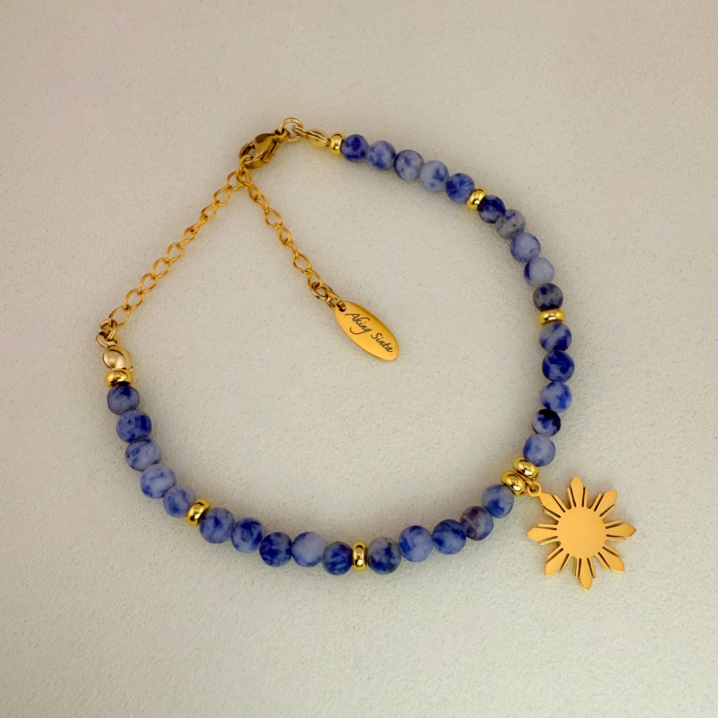 Aking Sinta Sodalite beaded bracelet featuring navy blue stones with white marbling and a gold 8-ray Araw charm; includes a gold extender chain with a teardrop-shaped metal tag engraved with "Aking Sinta" branding.