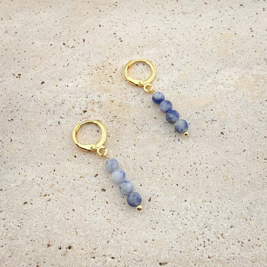 Pair of gold Aking Sinta huggie hoop earrings featuring rectangular deep blue Sodalite stone charms on a white marble background.