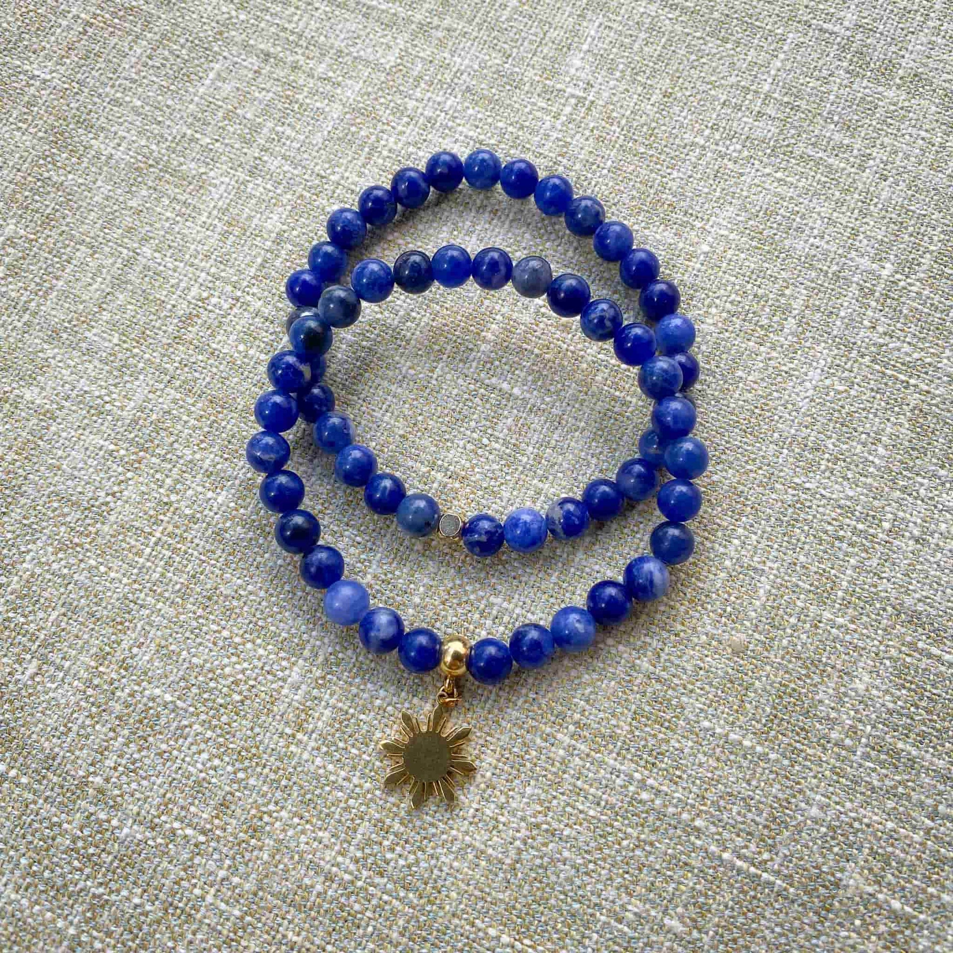 Two Aking Sinta Sodalite stretch bracelets stacked on a gray woven fabric background; one bracelet is a simple strand of blue stone beads, and the other features a gold Araw sun charm.