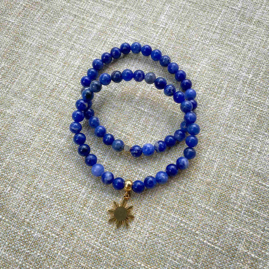 Two Aking Sinta Sodalite stretch bracelets stacked on a gray woven fabric background; one bracelet is a simple strand of blue stone beads, and the other features a gold Araw sun charm.