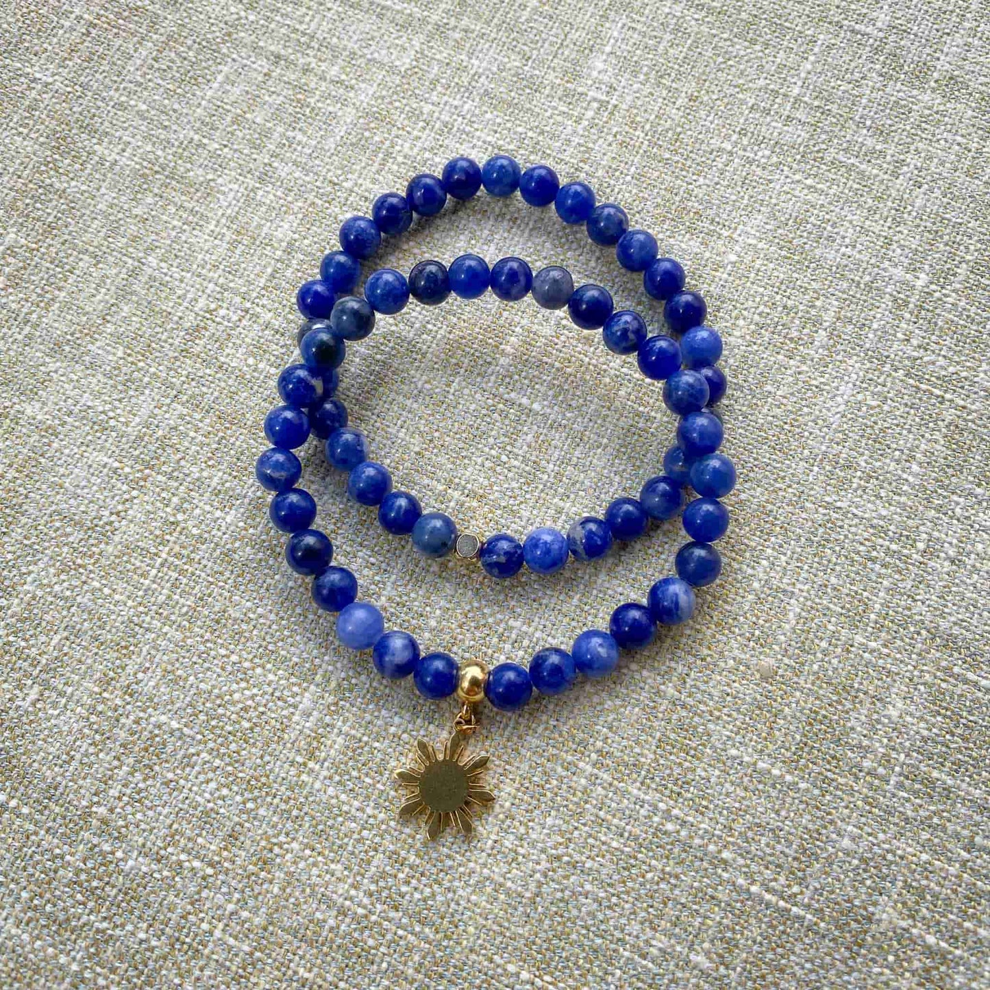 Two variants of the Sodalite blue stone stretch bracelet by Aking Sinta displayed together on a gray woven background; one plain minimalist strand and one featuring a gold Araw sun charm