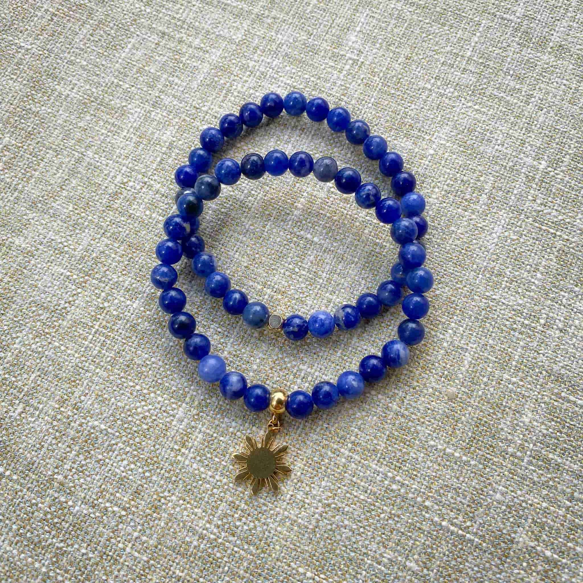 Two variants of the Sodalite blue stone stretch bracelet by Aking Sinta displayed together on a gray woven background; one plain minimalist strand and one featuring a gold Araw sun charm