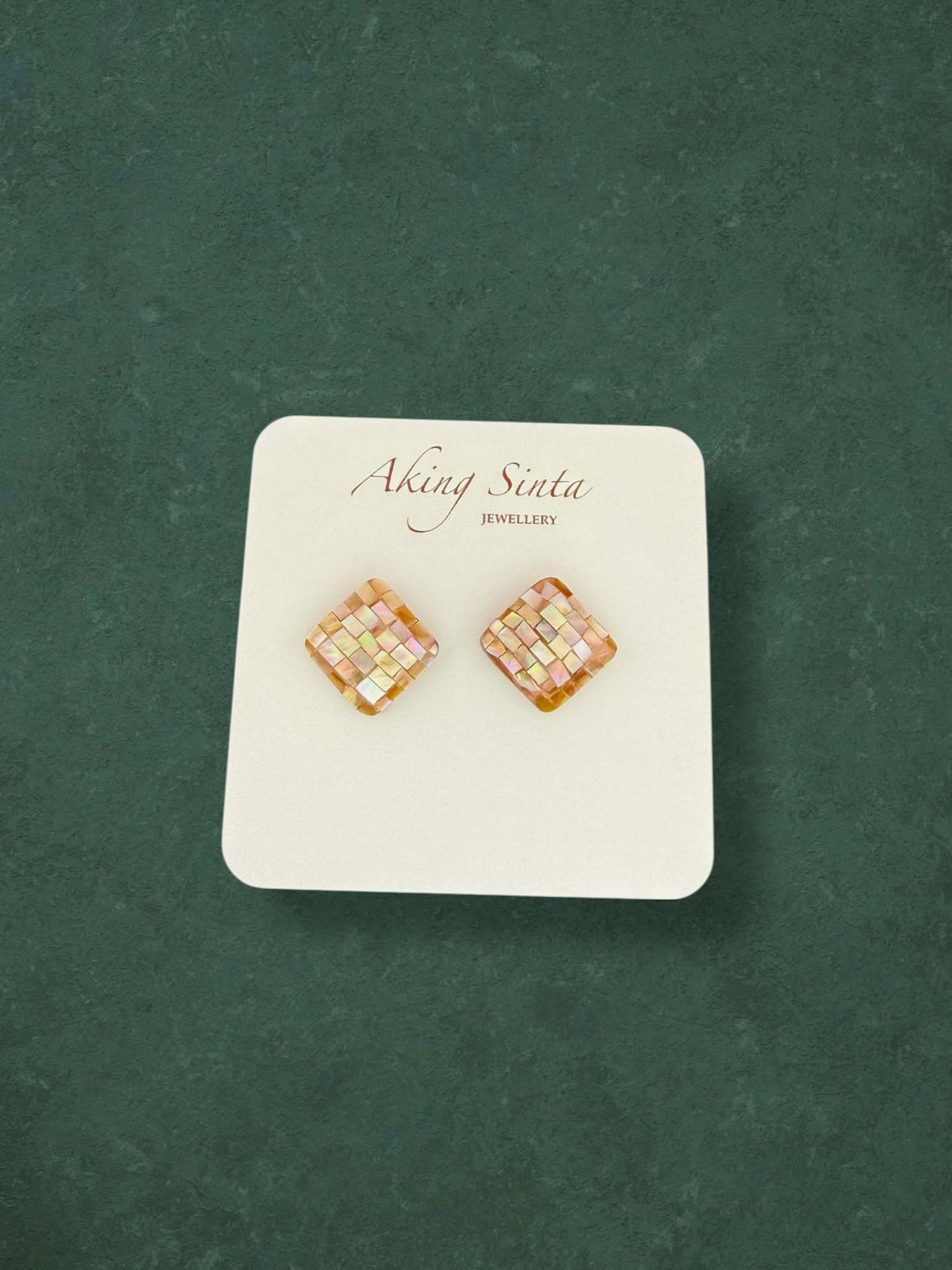 A pair of Aking Sinta square diamond-shaped stud earrings featuring a checkered mosaic mother of pearl inlay. The iridescent shell tiles display soft pink and gold shimmer on a white jewelry card