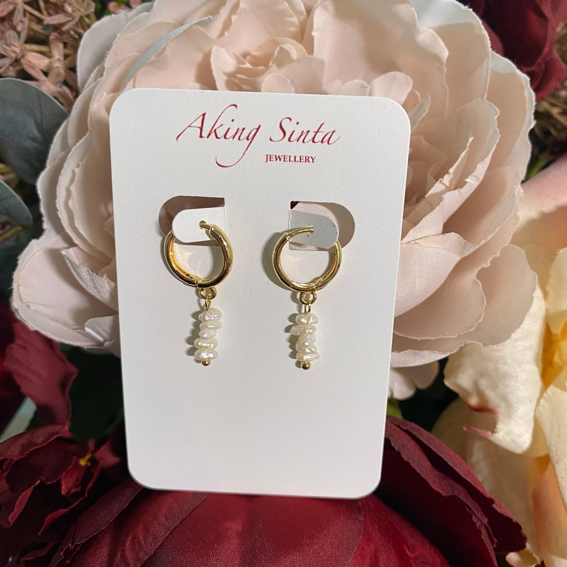 Aking Sinta Stacked Keshi Pearl Huggie earrings on a branded display card, resting on a bed of real pink and red roses.
