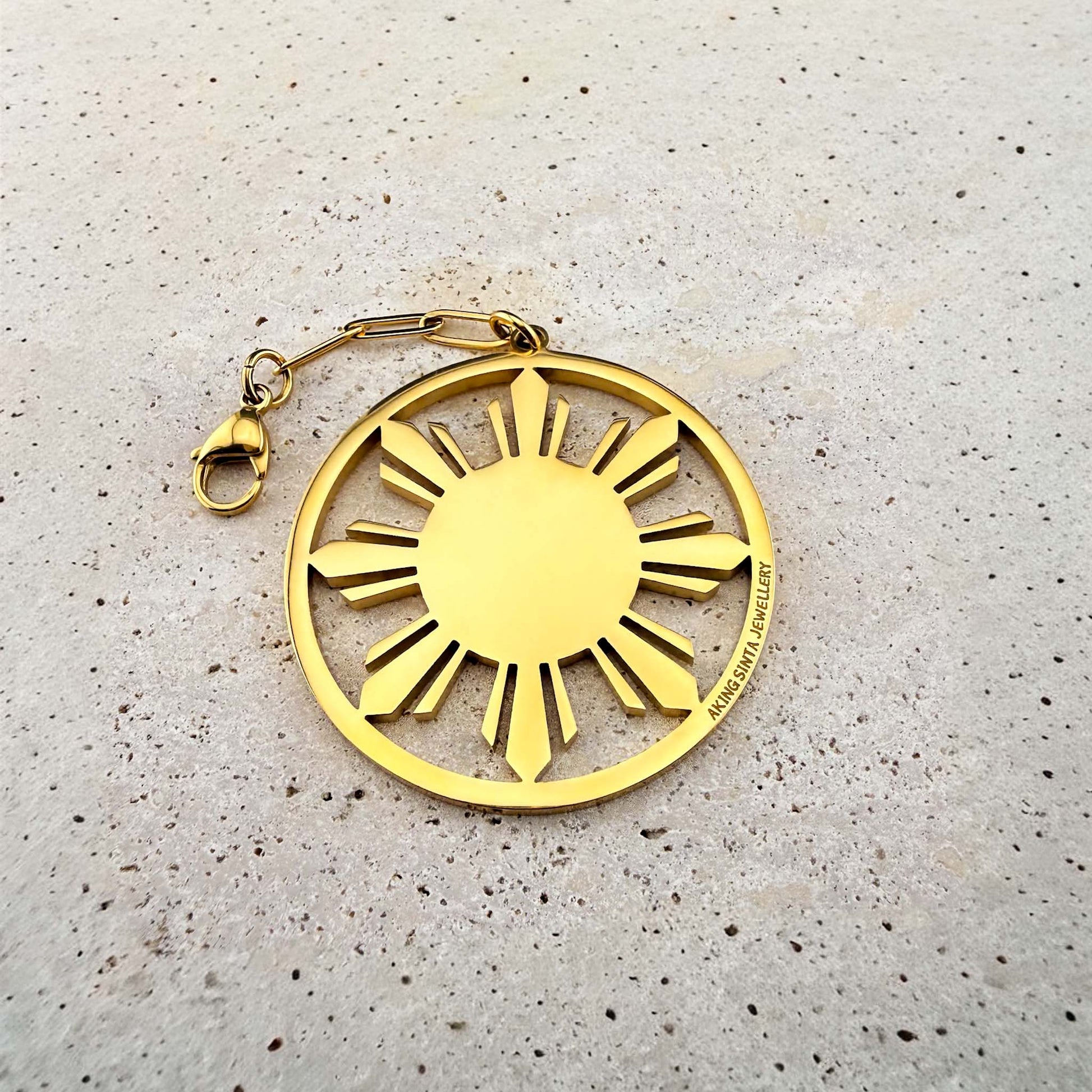 Close-up of the Aking Sinta gold Araw Sun bag charm resting on a white marble surface, showing the detailed medallion, chain, and clasp.