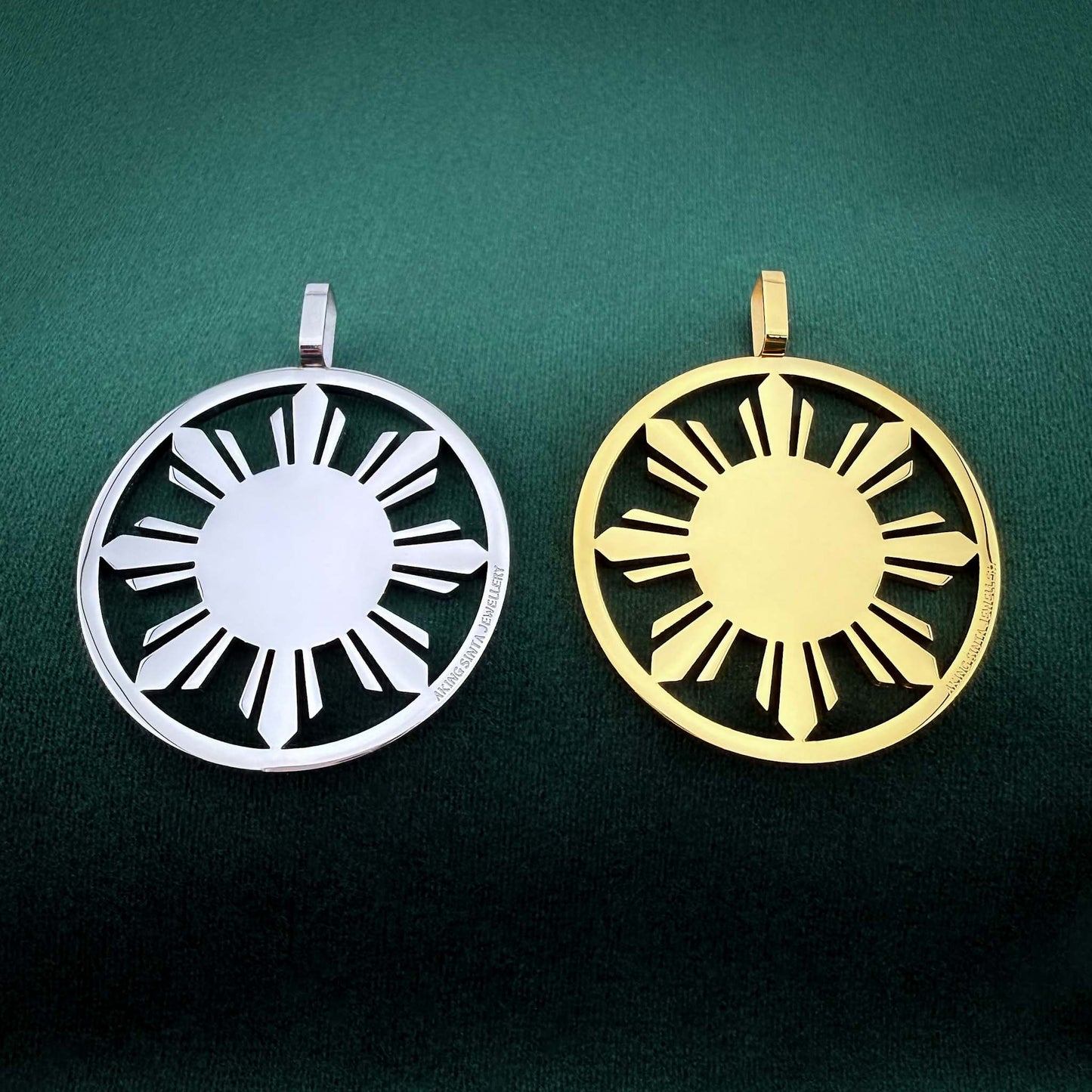 A pair of Aking Sinta statement Araw medallions, one in polished silver and one in gold, featuring a bold, sun-inspired design with a clean, cut-out aesthetic. The medallions are placed side-by-side on a rich, dark green velvet background, highlighting the sleek contrast between the two metallic finishes and the brand's minimalist, modern style