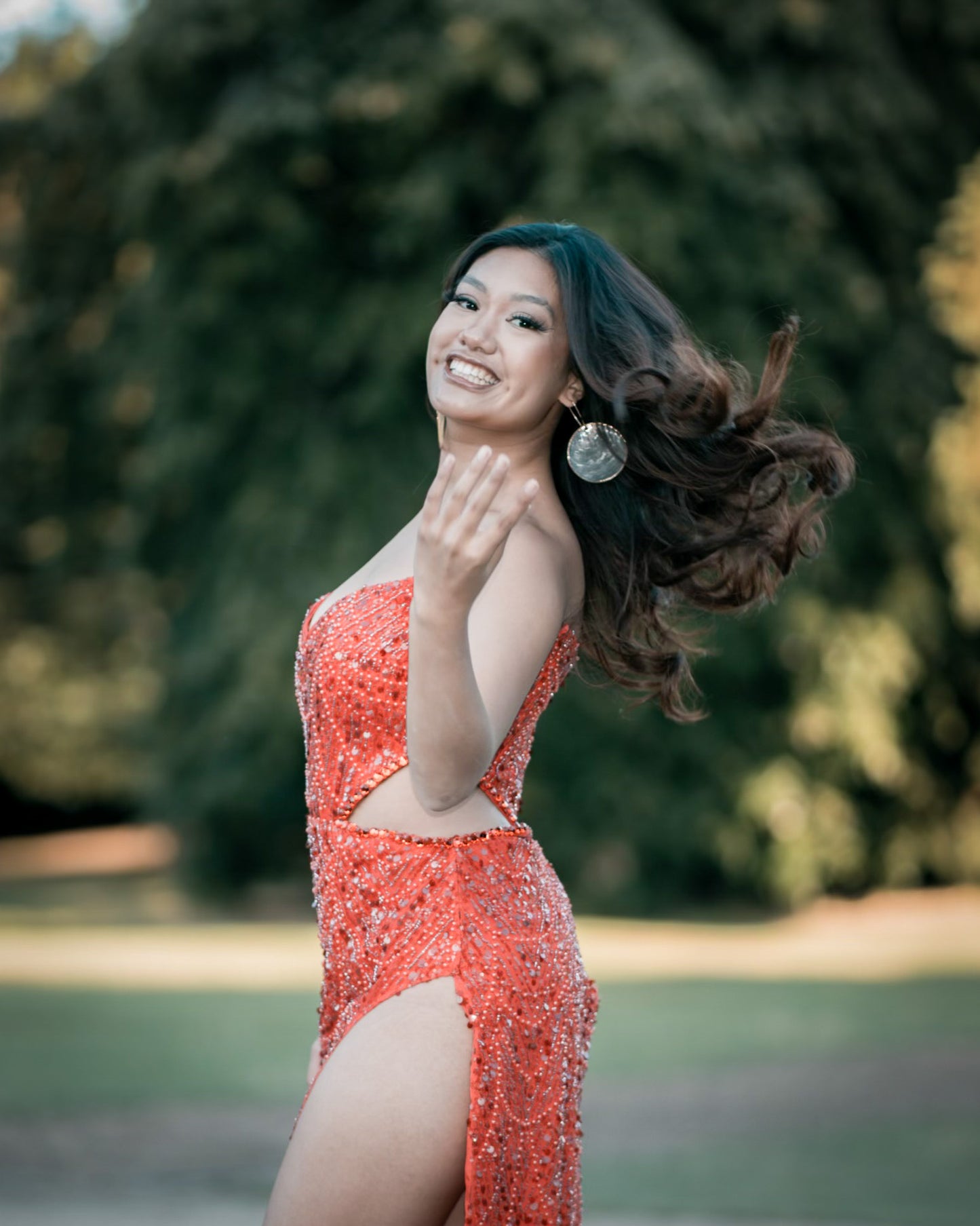 A woman with flowing hair wearing a vibrant red dress and large Aking Sinta Gold Lined Capiz Shell earrings; she is captured in a candid moment waving her hair, with the oversized iridescent white disks swinging gently and catching the light.
