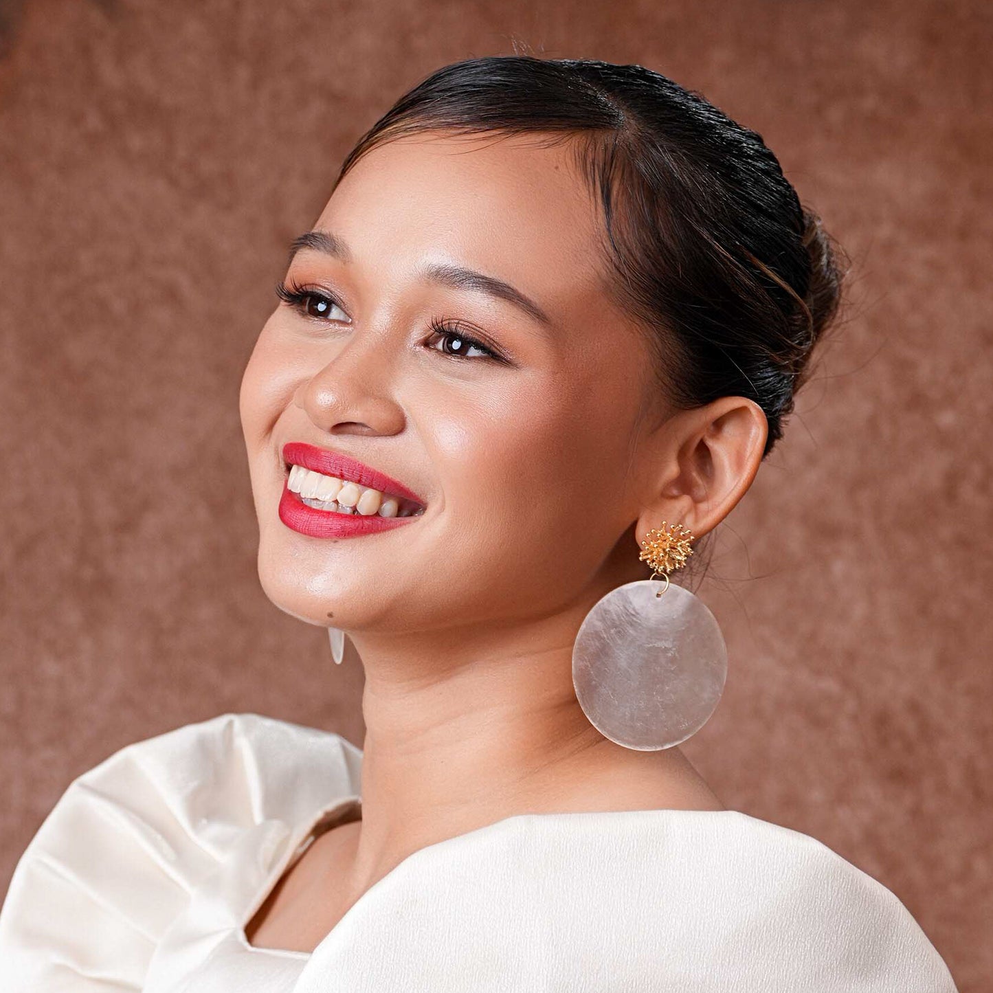A close-up of a model wearing Aking Sinta Statement Capiz and Coral Studs; featuring a large round iridescent white Capiz shell disk with a small, vibrant red coral bead at the center, sitting flush against the earlobe.