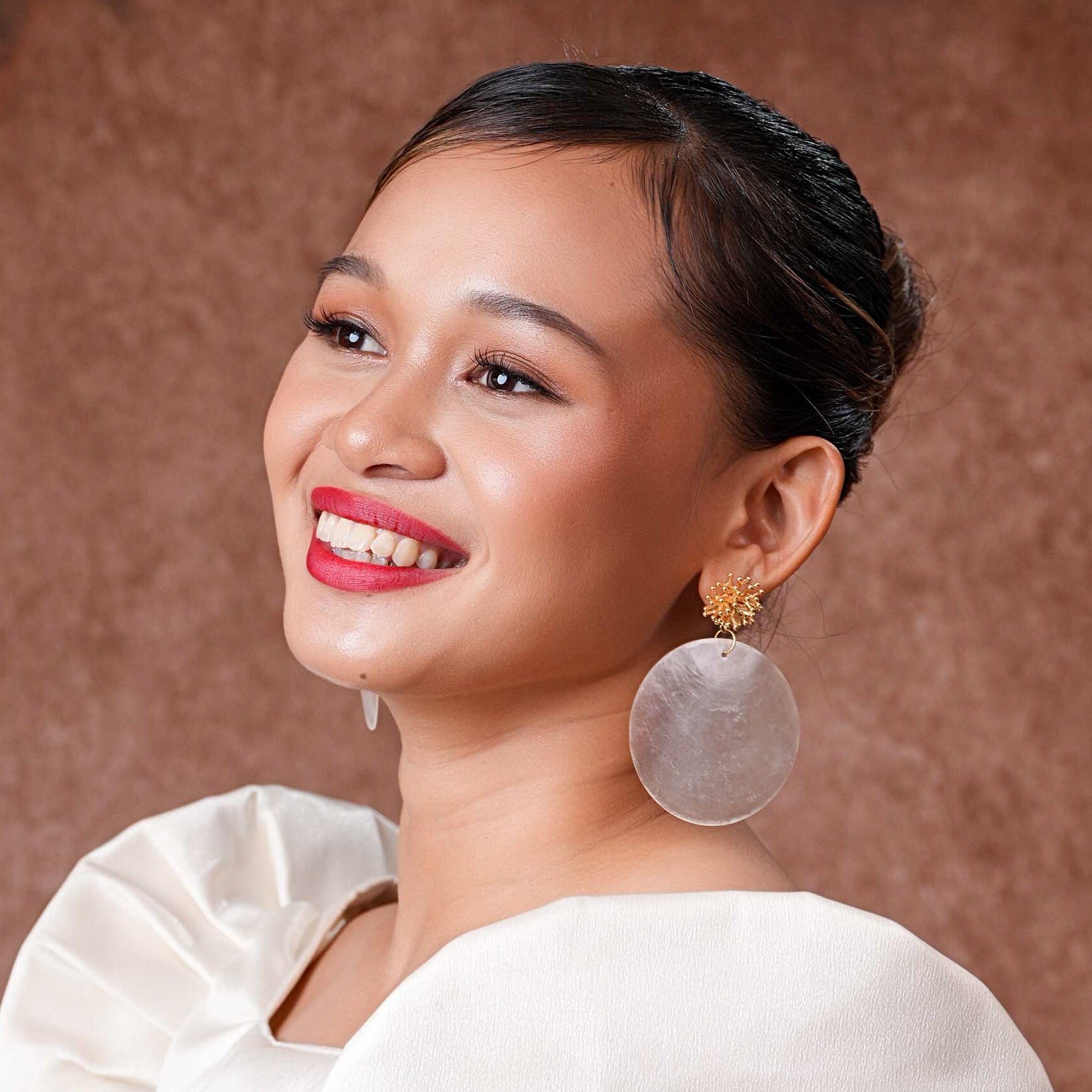 A close-up of a model wearing Aking Sinta Statement Capiz and Coral Studs; featuring a large round iridescent white Capiz shell disk with a small, vibrant red coral bead at the center, sitting flush against the earlobe.