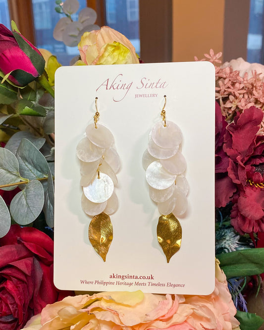 A pair of Aking Sinta Statement Cascading Capiz Shell Earrings mounted on a white branded cardboard display card; the long, tiered iridescent shell disks hang in a vertical row, set against a soft-focus background of real pink and white flowers.