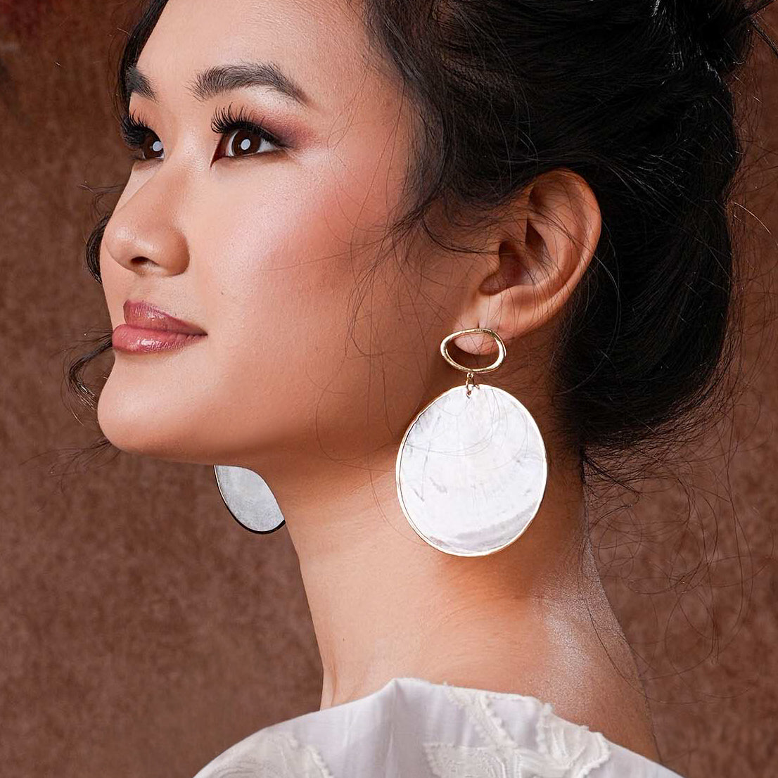 A close-up of a model wearing the Aking Sinta Statement Gold-Lined Capiz Shell Stud Earrings; the large, round iridescent white disk sits flat against the earlobe, framed by a thick, polished gold metal border.