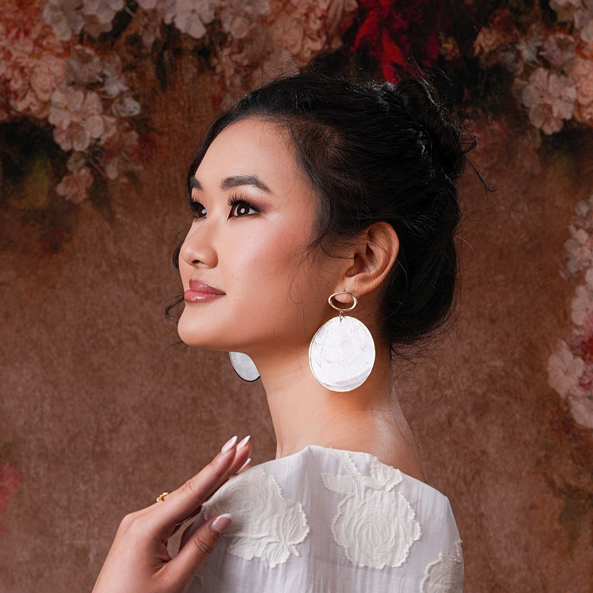 A close-up of a model wearing the Aking Sinta Statement Gold-Lined Capiz Shell Stud Earrings; the large, round iridescent white disk sits flat against the earlobe, framed by a thick, polished gold metal border.