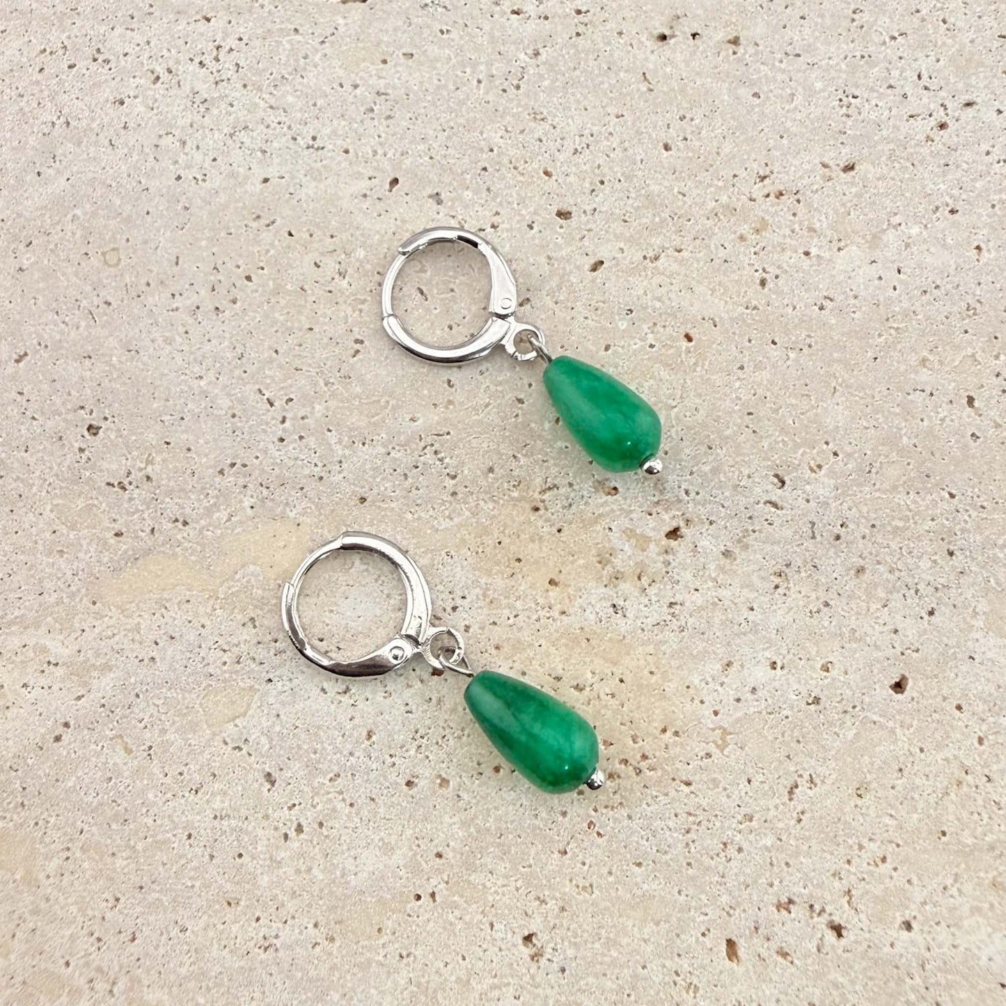 A pair of Aking Sinta silver huggie hoop earrings with dangling green jade teardrop charms. These minimalist gemstone drop earrings are pictured on a light-colored stone surface