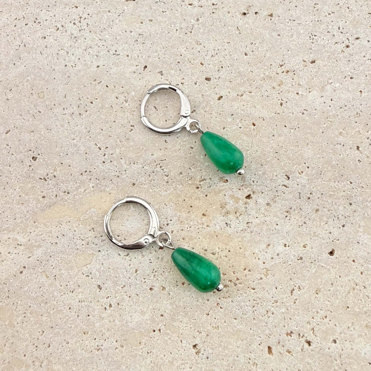 A pair of Aking Sinta silver huggie hoop earrings with dangling green jade teardrop charms. These minimalist gemstone drop earrings are pictured on a light-colored stone surface