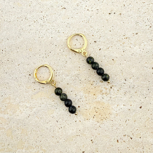 A pair of gold huggie earrings on a white background, each featuring a vertical drop of three small, round Serpentine beads in a light chartreuse green, hanging from a polished gold hoop.