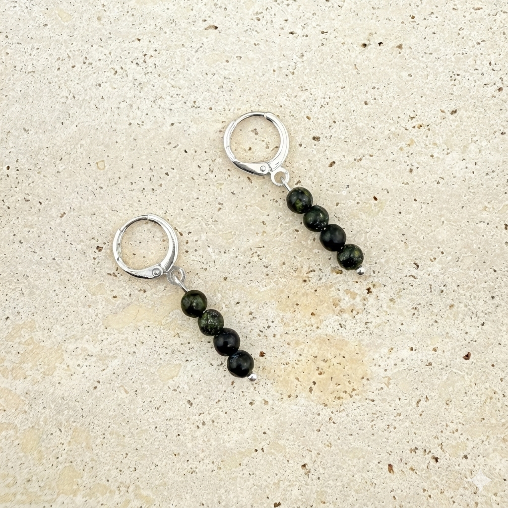 A pair of Aking Sinta silver huggie earrings on a textured white rock background; each earring is securely closed and features a vertical drop of three translucent, light-green Serpentine beads hanging from a polished silver hinged hoop.