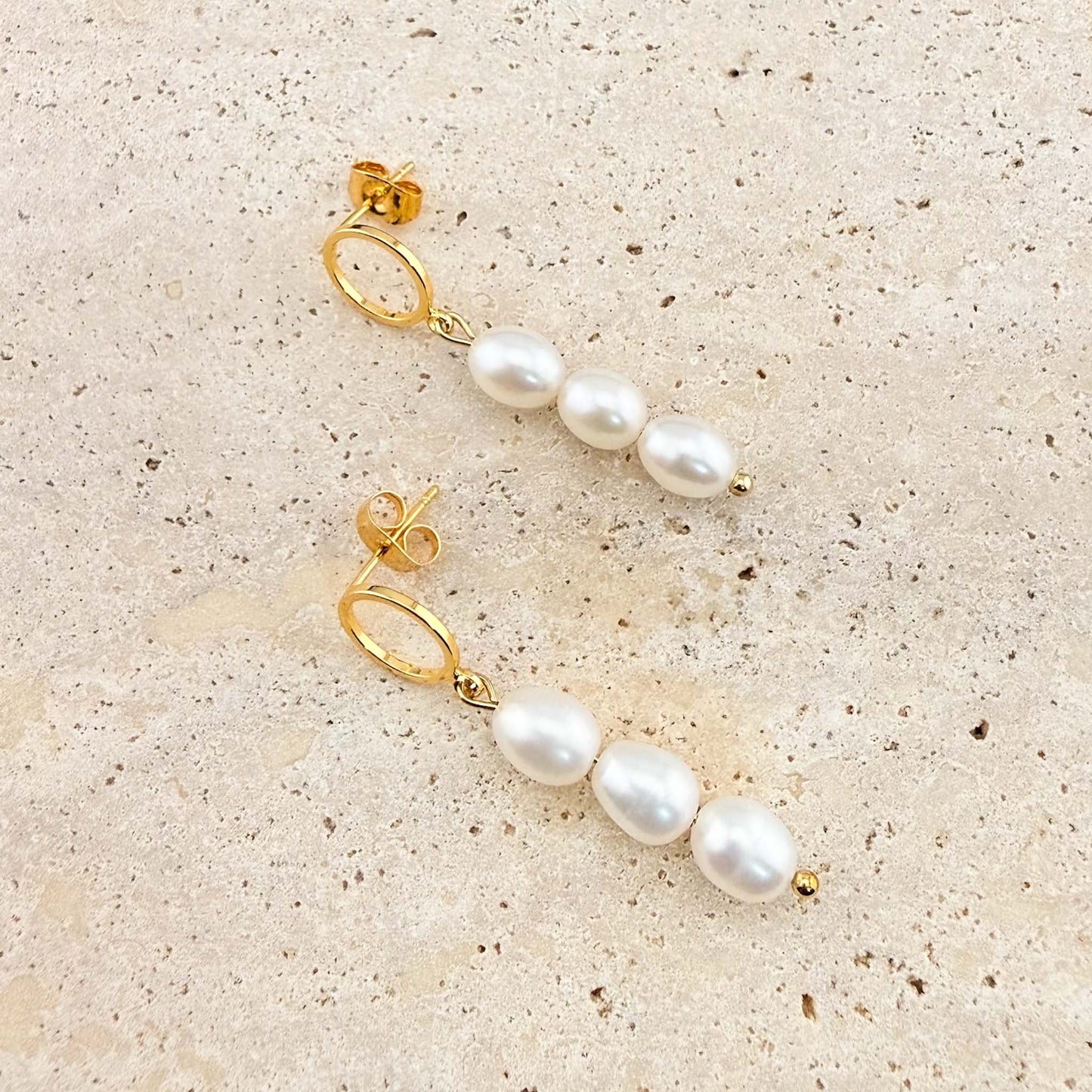 A pair of Aking Sinta gold triple pearl drop earrings. Each earring features three white freshwater pearls suspended from a polished gold circle stud, laid out on a natural stone surface